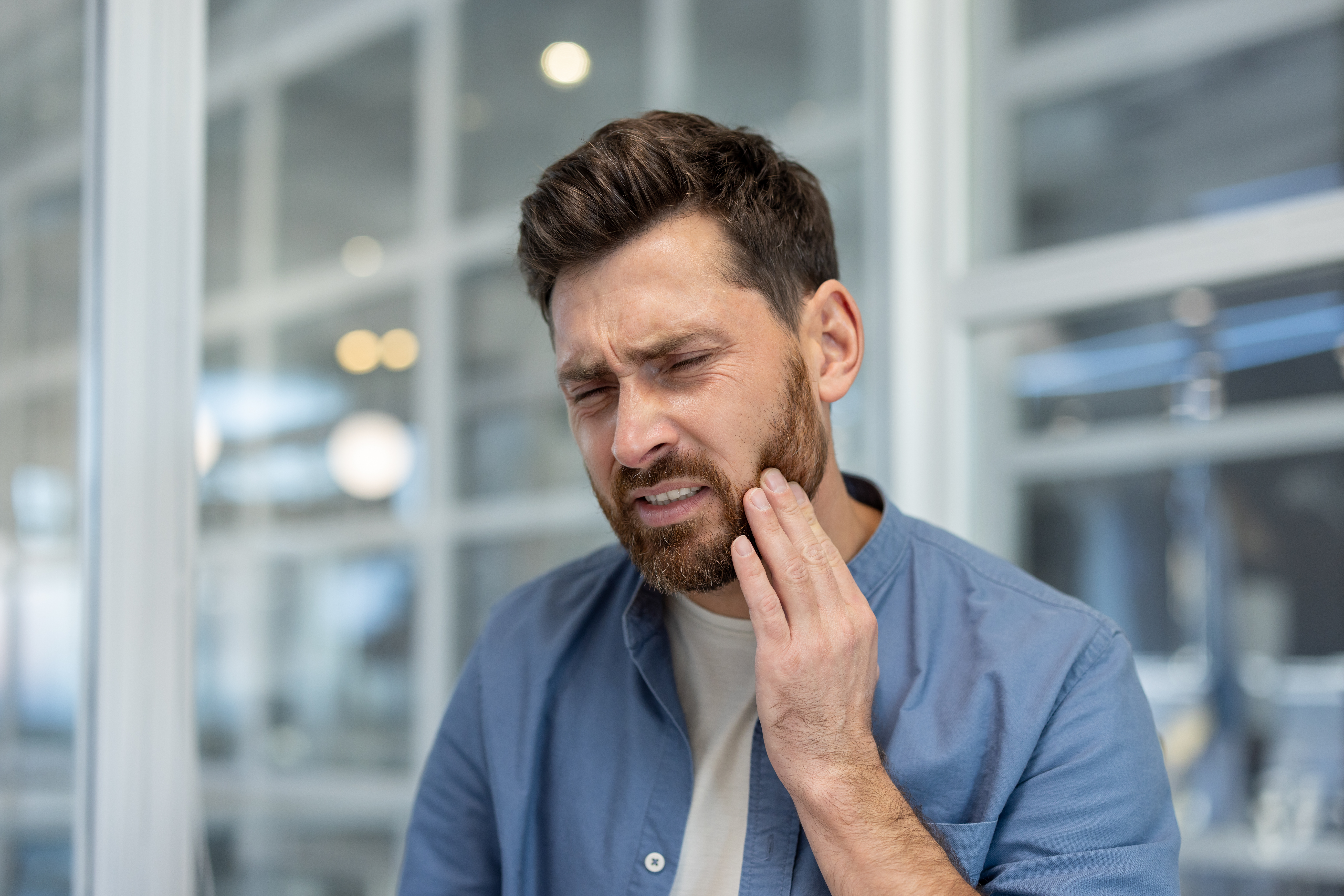 Man with beard experiencing sharp tooth pain, holding cheek with hand, frowning, expressing discomfort from a dental problem or oral health discomfort